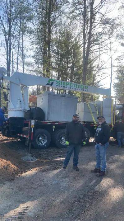 Two workers in a dirt trench install PVC pipes onto a concrete septic tank lid, using a power drill and level.