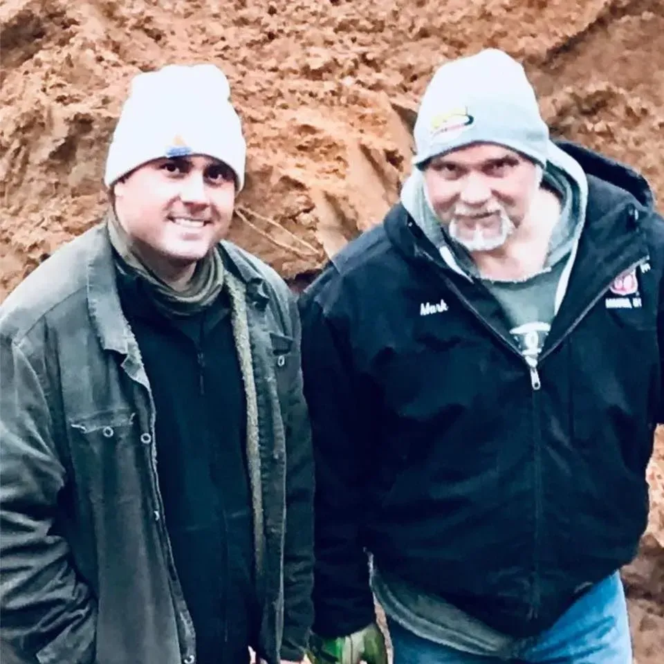 A person in a dark green polo and black gloves smiles while holding papers in a bright, plant-filled office.