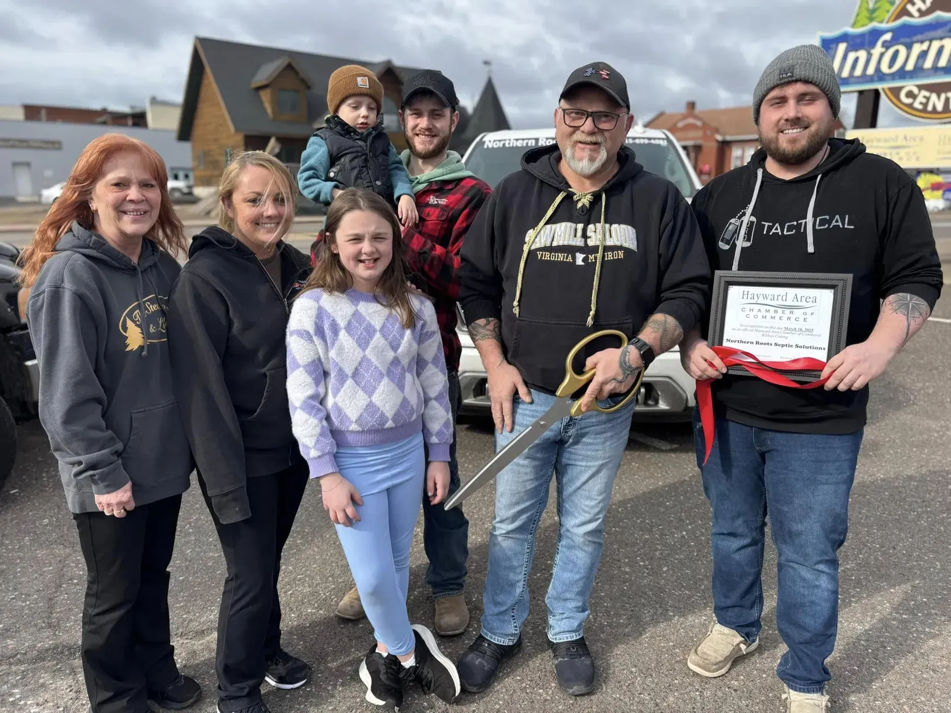 A smiling person in a dark green polo and black gloves holds papers while standing in a bright, plant-filled indoor space.