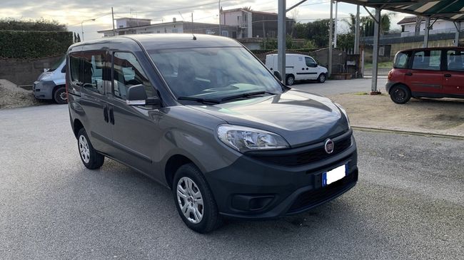 Un'auto compatta grigia parcheggiata su un piazzale ghiaioso, con altri veicoli e una recinzione sullo sfondo.