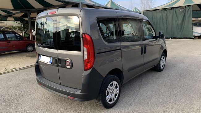 Furgone compatto grigio parcheggiato all'aperto sotto delle tende in un piazzale ghiaioso, vista posteriore a tre quarti.