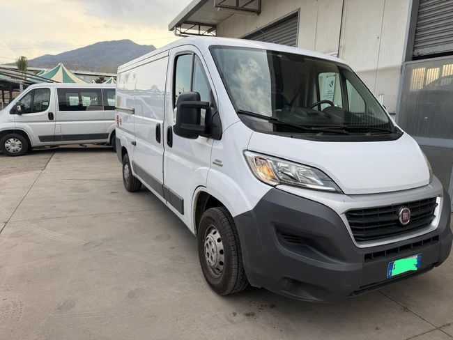 Un furgone bianco per il trasporto merci è parcheggiato in un piazzale accanto a un edificio, con un altro furgone sullo sfondo.