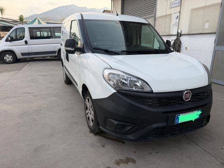 Furgone bianco parcheggiato in un piazzale, vista frontale con montagne e un altro furgone sullo sfondo.