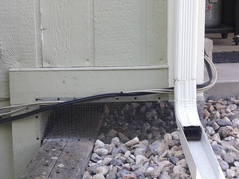 Exterior house wall with wire mesh over a vent, next to a downspout and gravel.