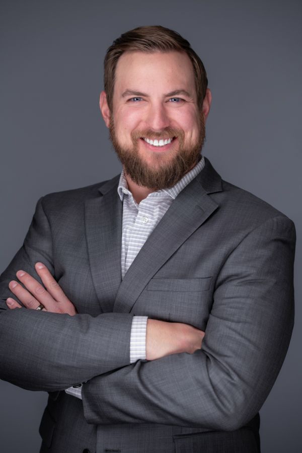 A man with a beard is wearing a suit and smiling with his arms crossed.