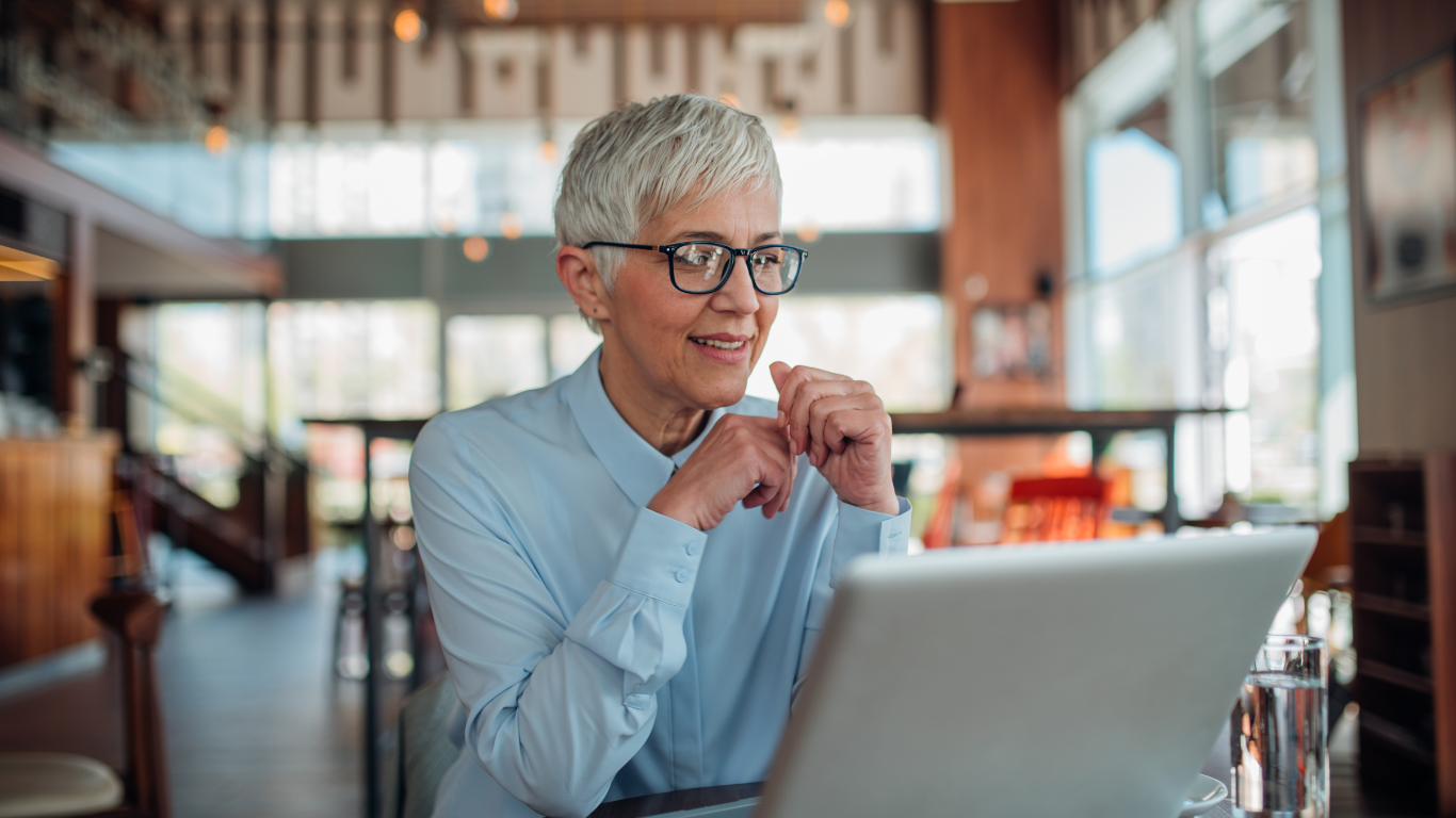An older woman is sitting at a table with designer eyeglasses using a laptop.