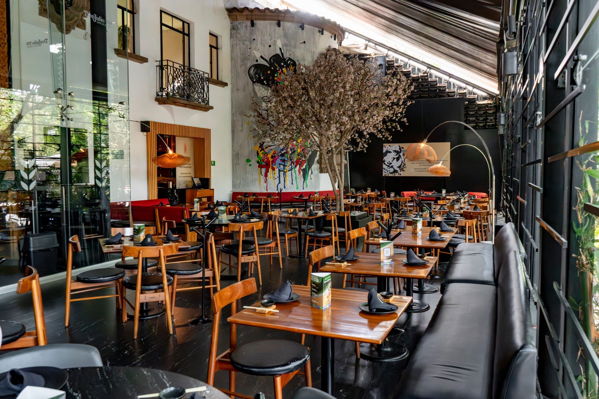 Interior de restaurante moderno con mesas de madera, sillas negras y altos árboles de interior bajo un techo alto.