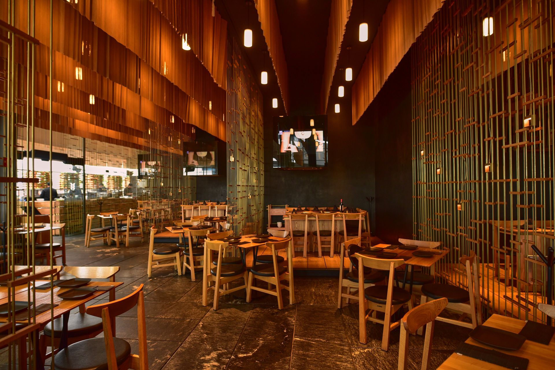 Interior de restaurante moderno con iluminación cálida, mesas y sillas de madera y lámparas colgantes.