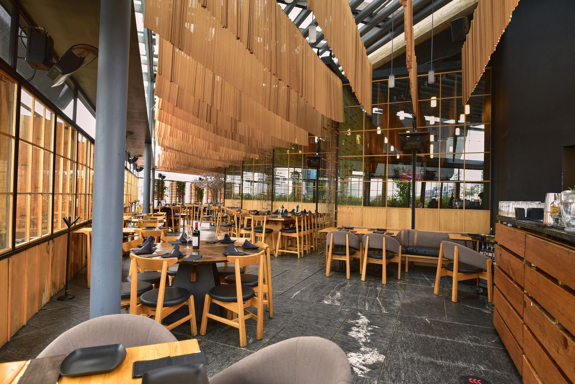 Interior de restaurante moderno con mesas de madera, listones de madera colgantes y grandes ventanales.