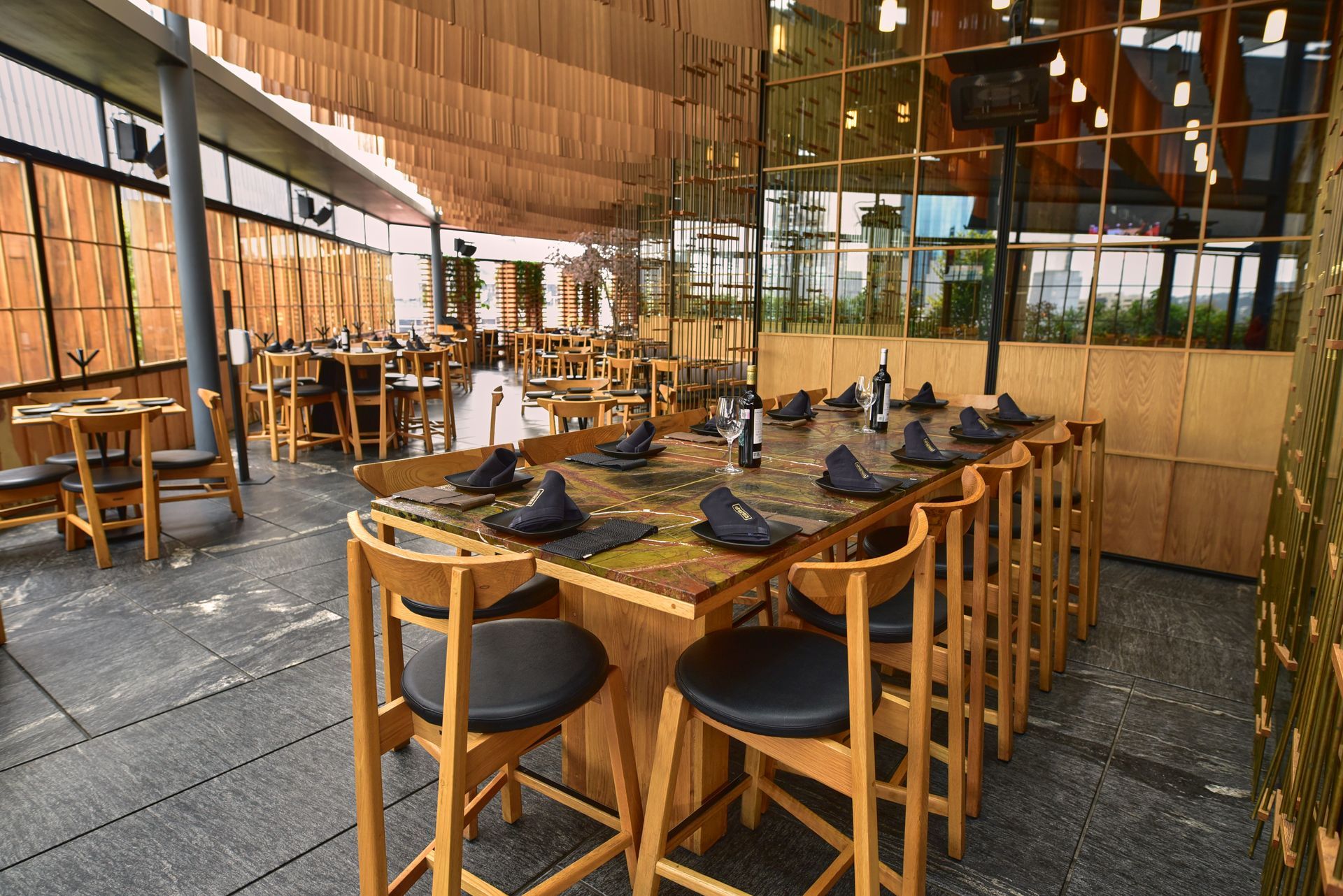 Terraza de cafetería con mesas y taburetes de madera bajo un toldo, situada junto a paredes de cristal.