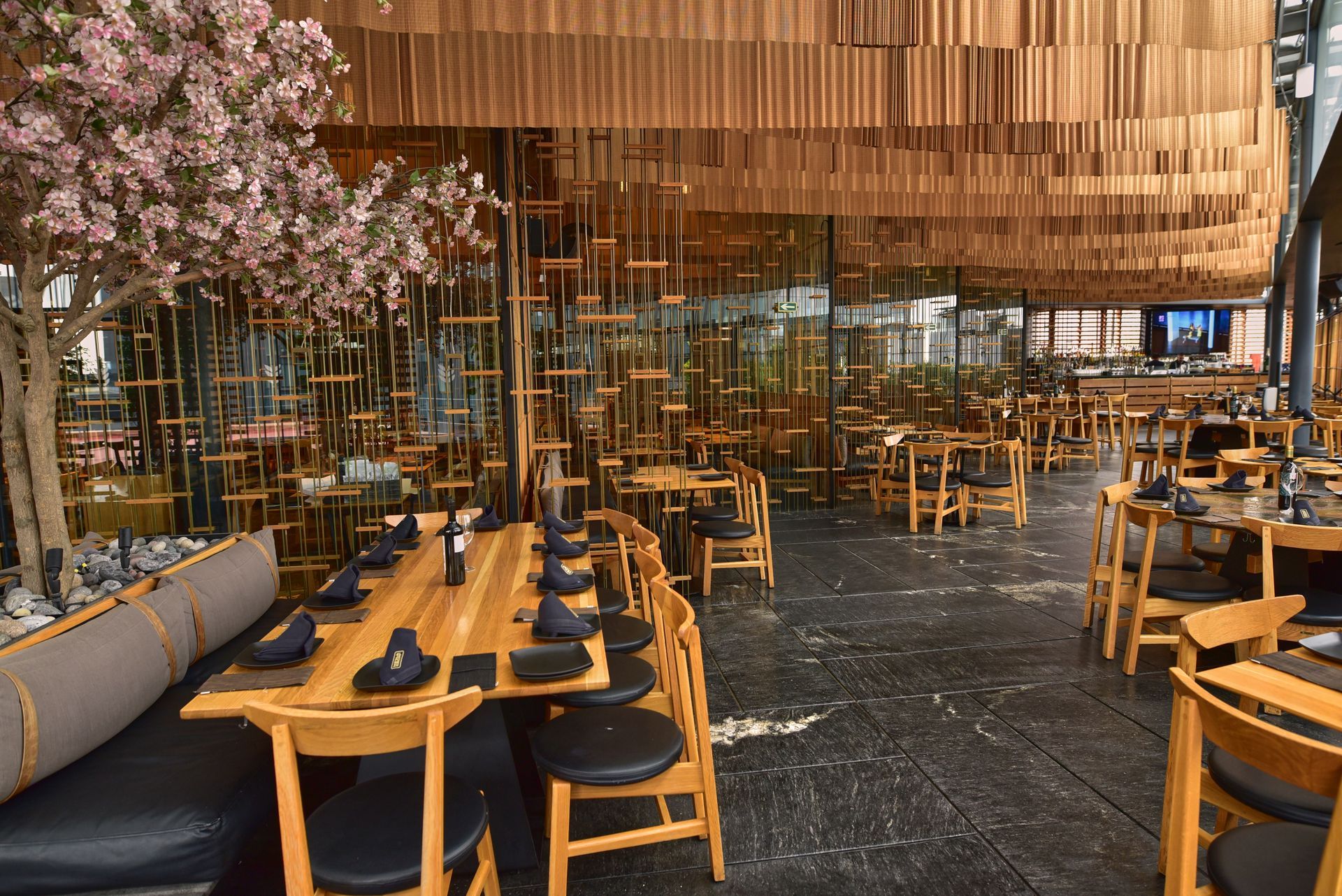 Interior de restaurante moderno con mesas y sillas de madera bajo un cálido techo de madera y decoración floral.