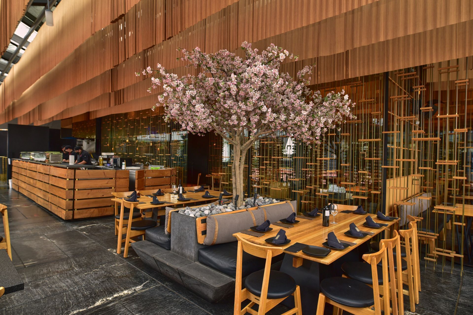 Terraza exterior del restaurante con mesas de madera, sillas negras y un árbol rosa en flor bajo una fachada de cobre.
