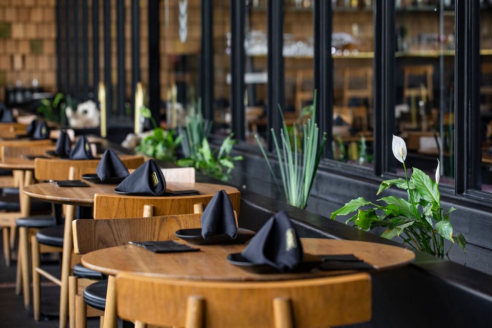 Comedor vacío de un restaurante con mesas de madera, servilletas negras y plantas verdes a lo largo de una pared con ventana.