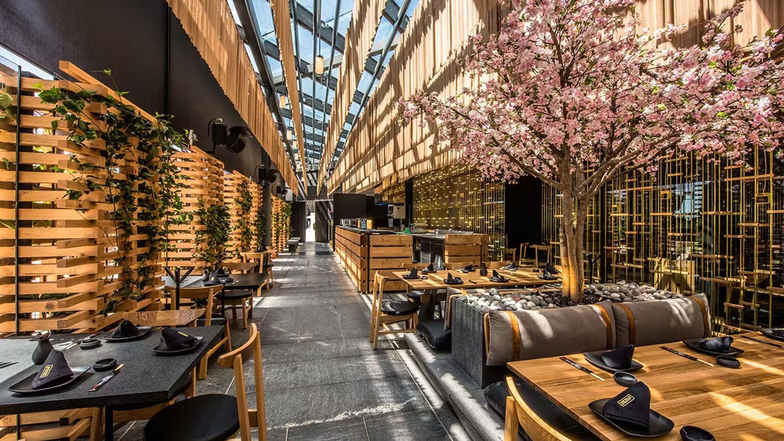 Elegante salón interior con asientos de madera, paredes de bambú y árboles de flores rosas bajo un largo techo con tragaluz.