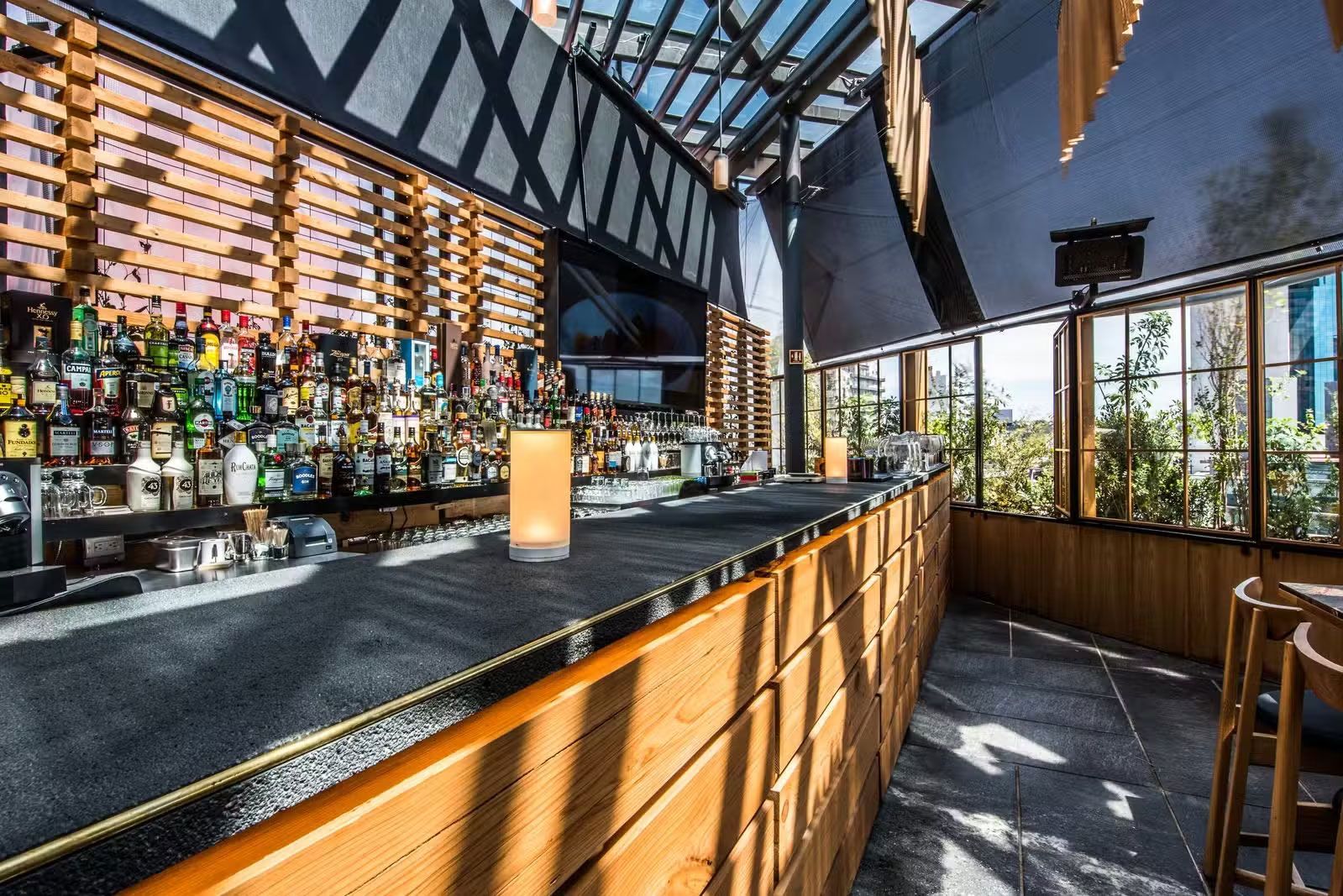 Bar moderno y luminoso con barra de madera, botellas y grandes ventanales con vistas a la vegetación.
