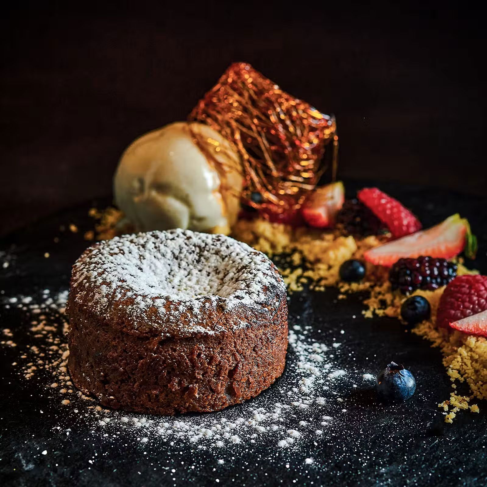 Pastel de chocolate espolvoreado con azúcar glas, cubierto con helado, en un plato de postre oscuro con frutos rojos.