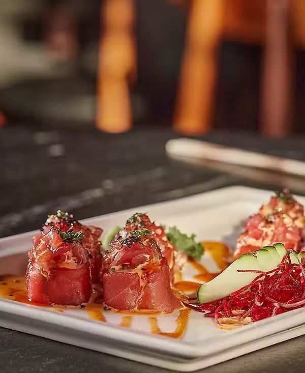 Rollos de sushi emplatados con salmón y aguacate en un plato blanco, rociados con salsa en una mesa oscura.