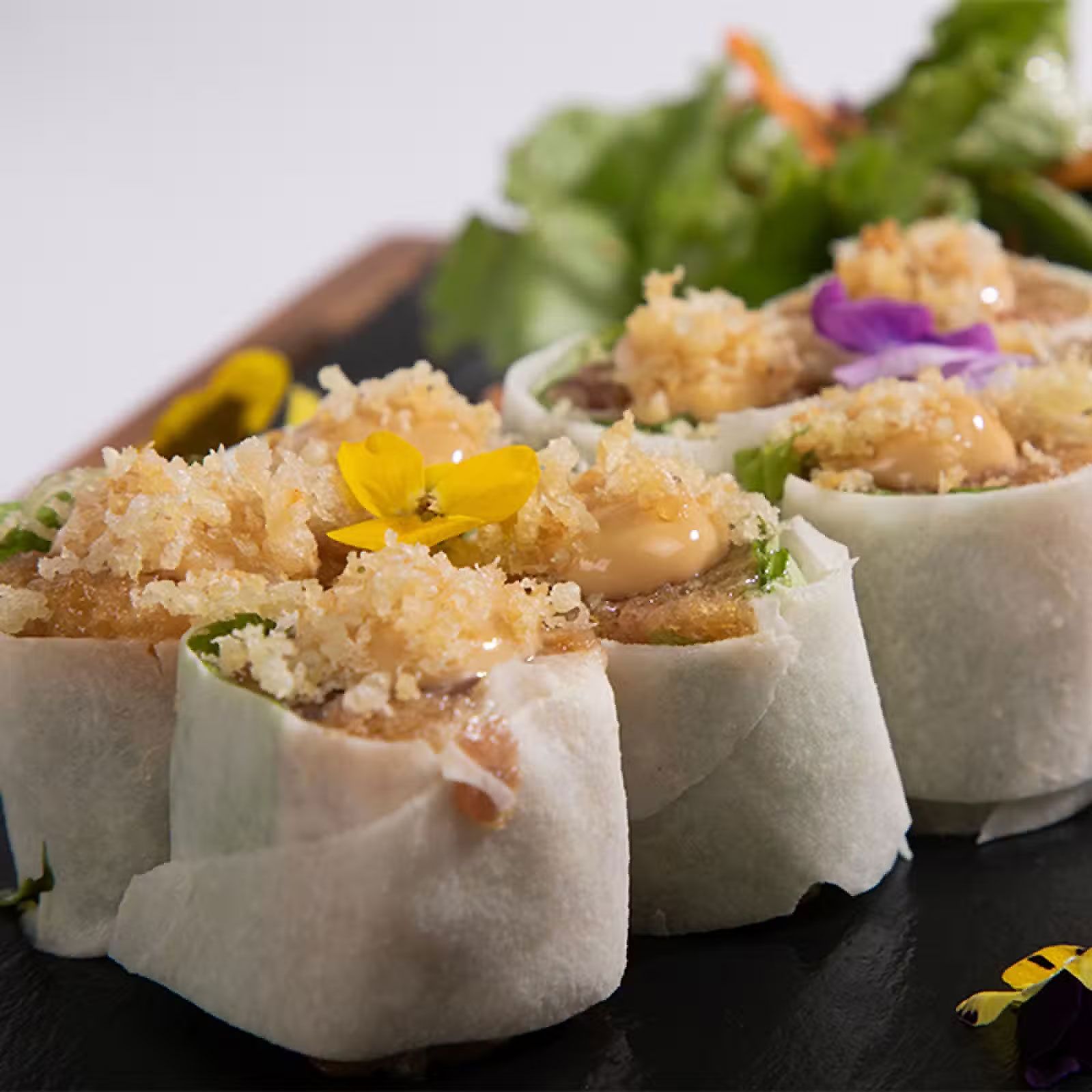 Postres pequeños decorados con flores y cubiertos con migas, servidos en una bandeja negra.