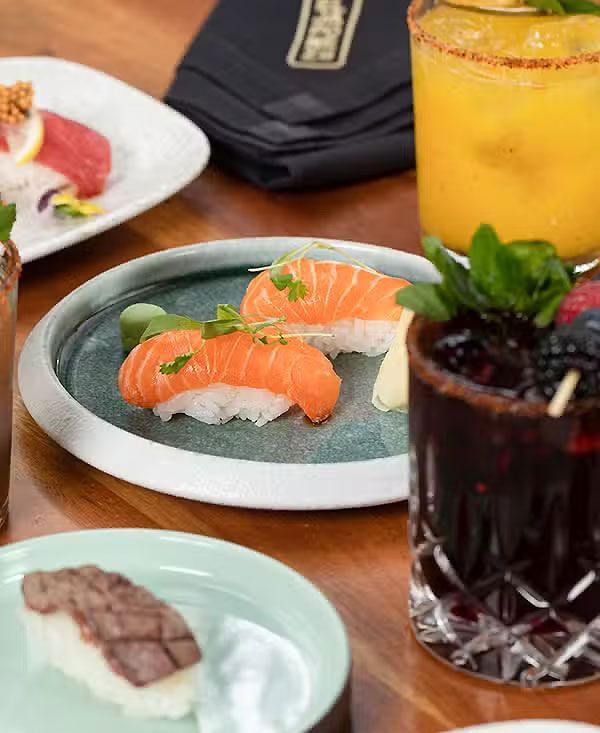Platos de sushi y postre sobre la mesa de un restaurante, acompañados de coloridos cócteles.