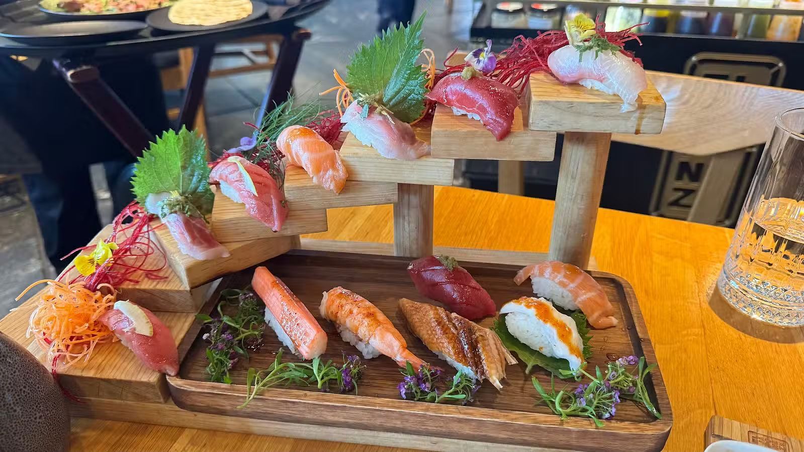 Plato de sushi sobre una tabla de madera con coloridos nigiri y guarniciones, servido en una mesa de restaurante.