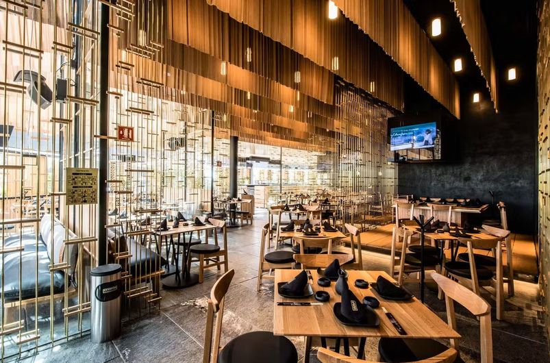 Interior de restaurante moderno con mesas de madera, lámparas colgantes e iluminación ambiental cálida.
