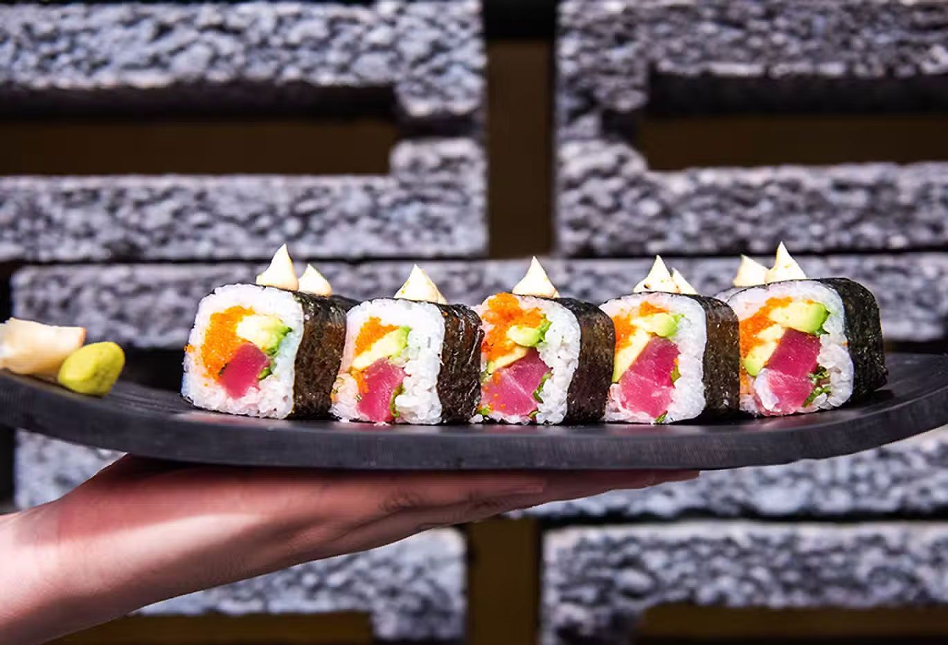 Rebanadas de rollo de sushi con arroz blanco, relleno rosa y guarnición verde en un plato negro, sostenidas en una mano.
