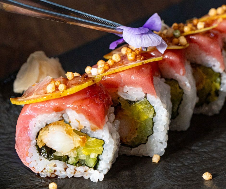 Rollos de sushi gourmet cubiertos con pescado rosado, semillas de sésamo y una flor comestible morada sobre un plato oscuro.