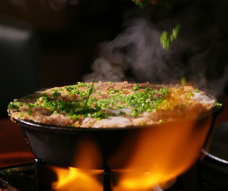 Sartén humeante con comida dorada adornada con hierbas picadas sobre fuego directo.
