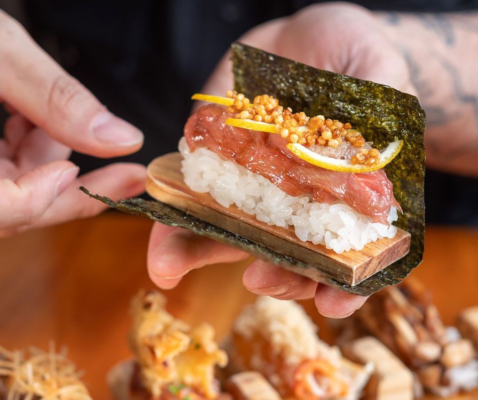 Manos sosteniendo un rollo de sushi con arroz, atún y guarnición de naranja sobre láminas de nori.