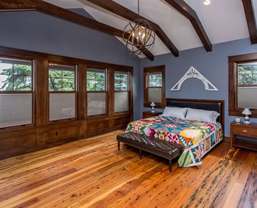 Bedroom with blue walls, wooden floors and trim, a bed with a colorful quilt, and large windows.
