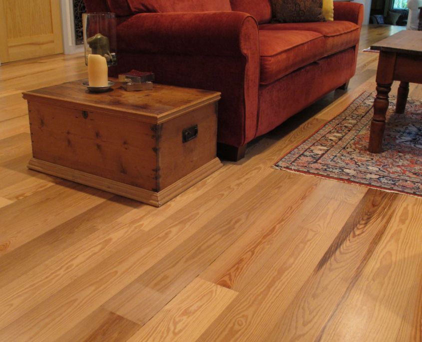 Wooden floor in a living room; a red couch, wooden chest, and a small rug are present.
