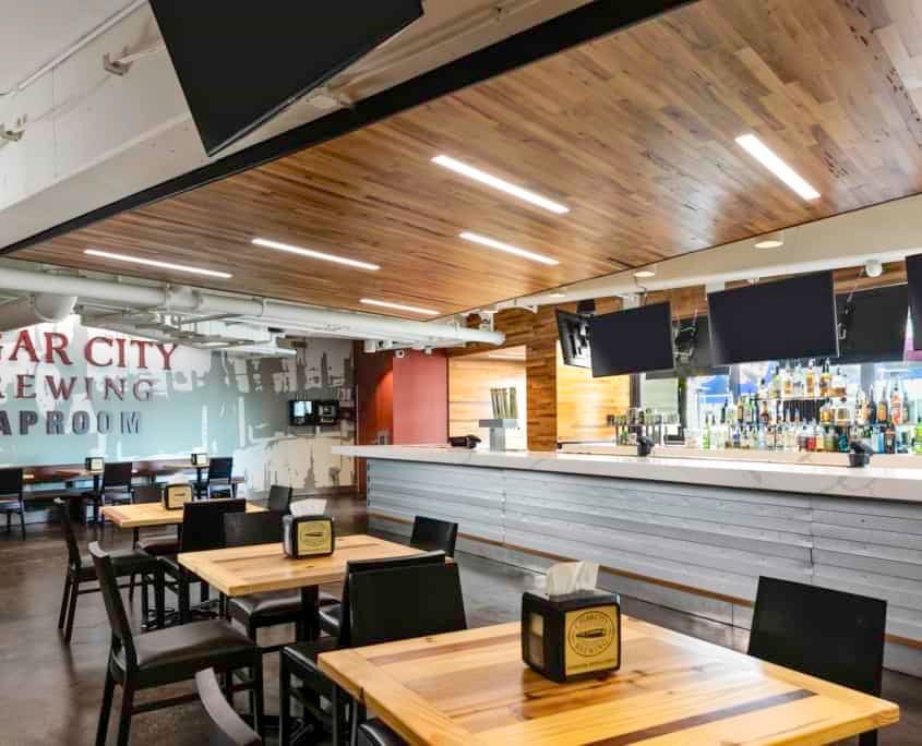Interior of Sugar City Brewing taproom with tables, bar, and TVs. Wooden ceiling, white bar, and gray tables.