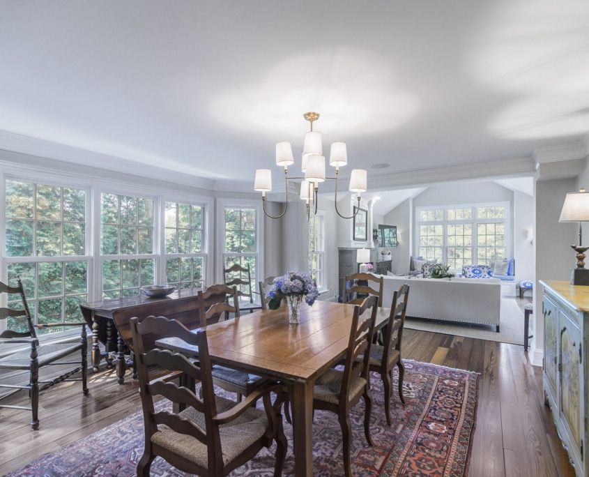 Dining room with wooden tables and chairs, chandelier, rug, and windows overlooking greenery.