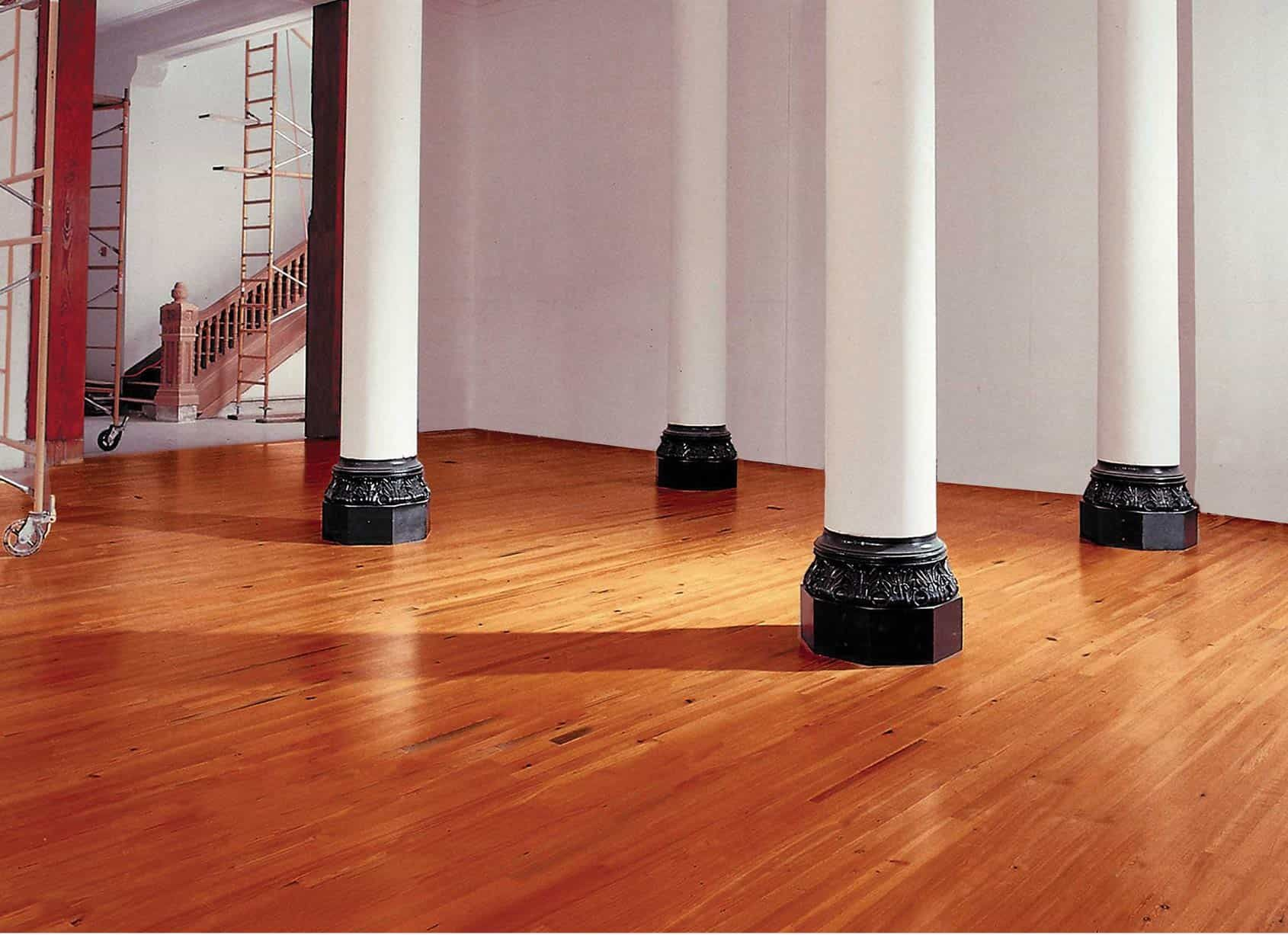 Interior of a room with polished wooden floor, white pillars, and unfinished walls; visible staircase in background.