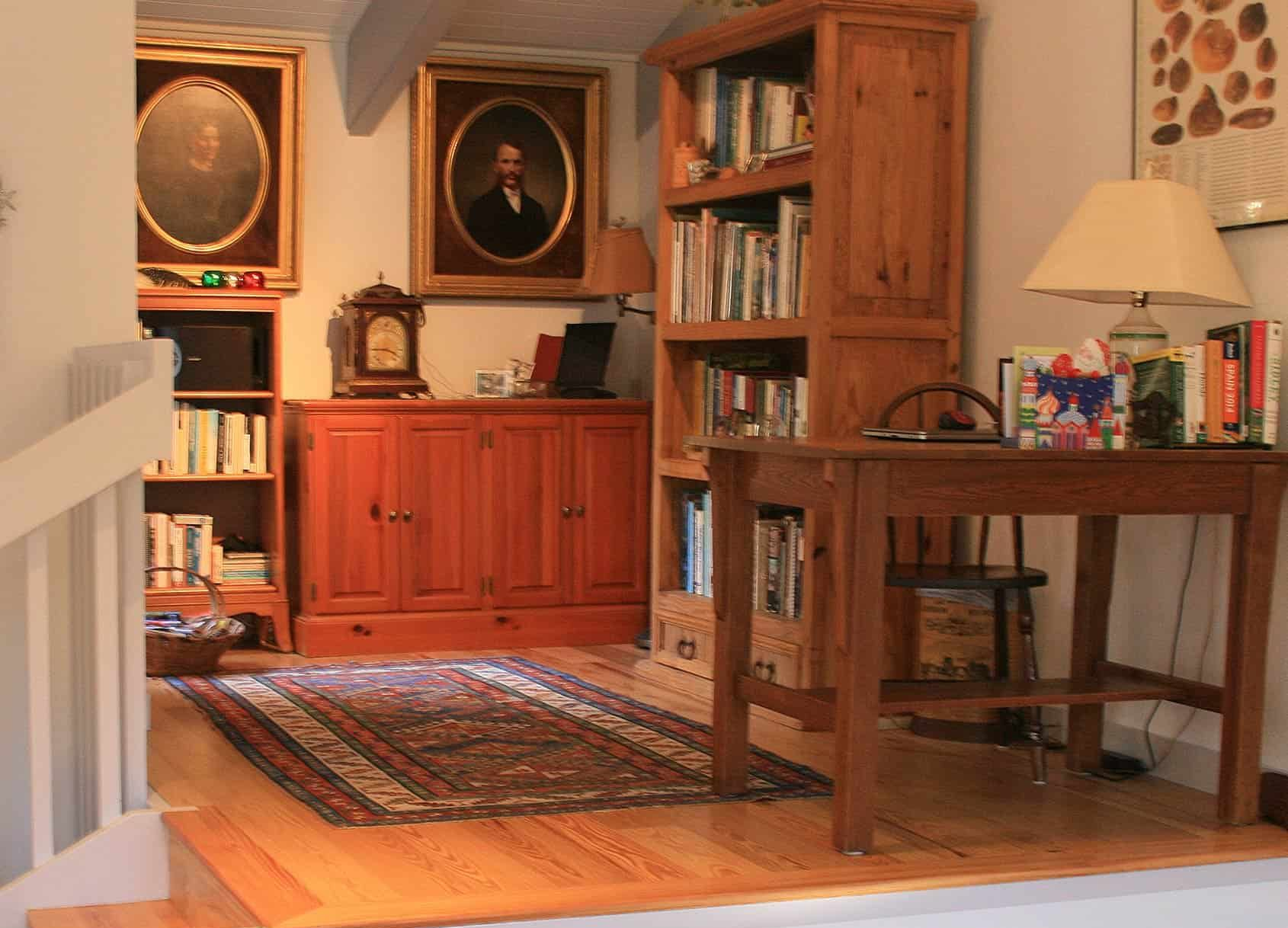 Cozy study with wood furniture, bookshelves, rug, portraits, and desk with lamp.
