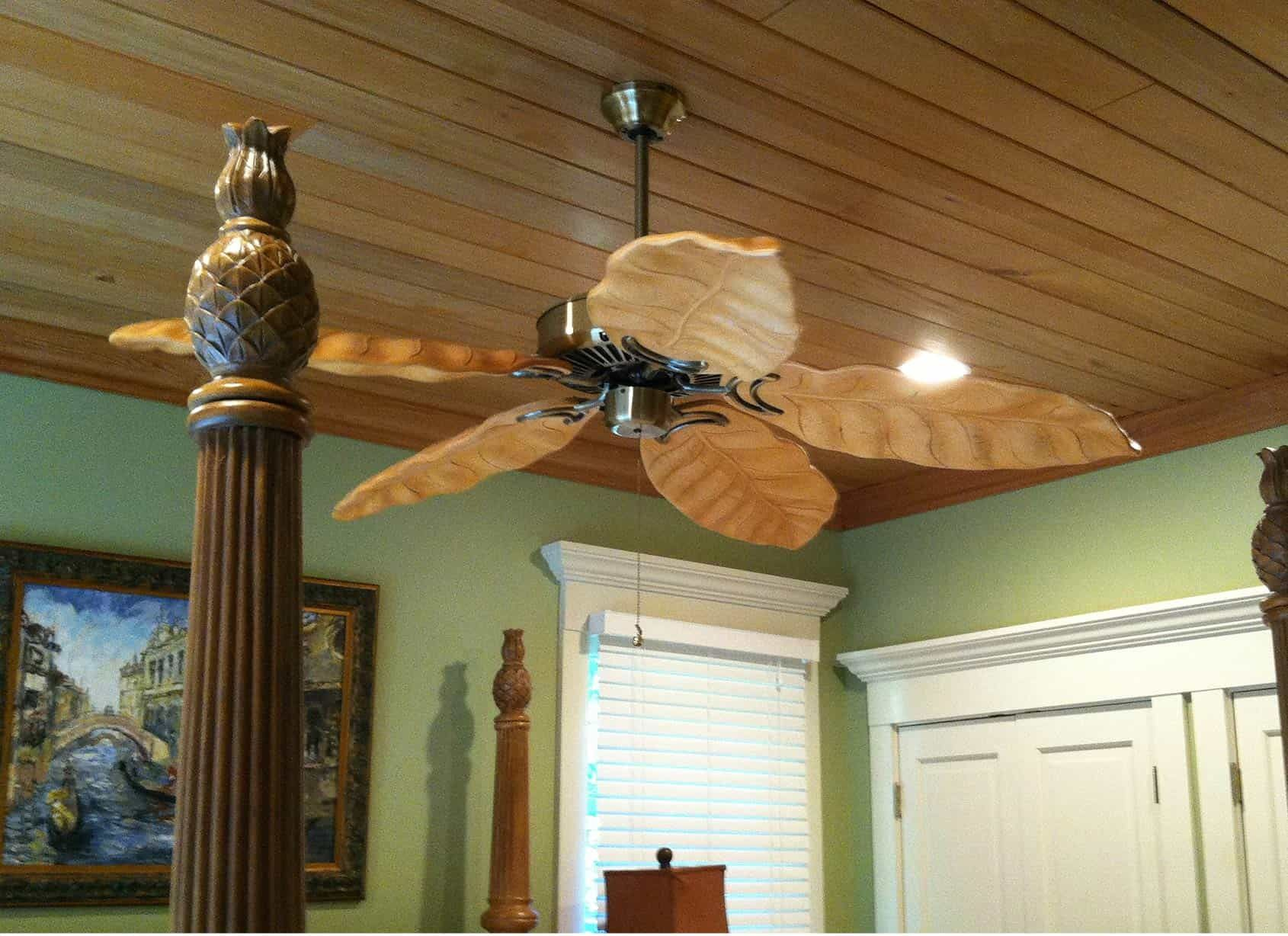 Ceiling fan with wooden blades, mounted on wood paneled ceiling, next to bedpost with pineapple top.