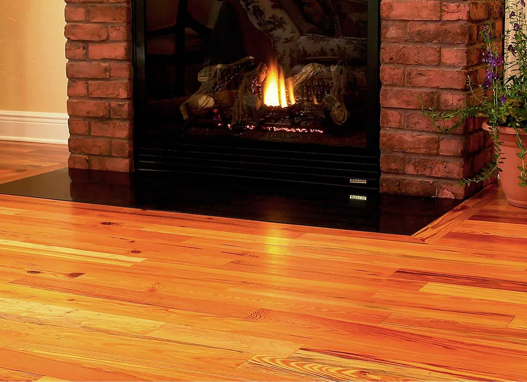 Fireplace with burning flames, set in a brick frame, on hardwood floors, with a potted plant on the right.