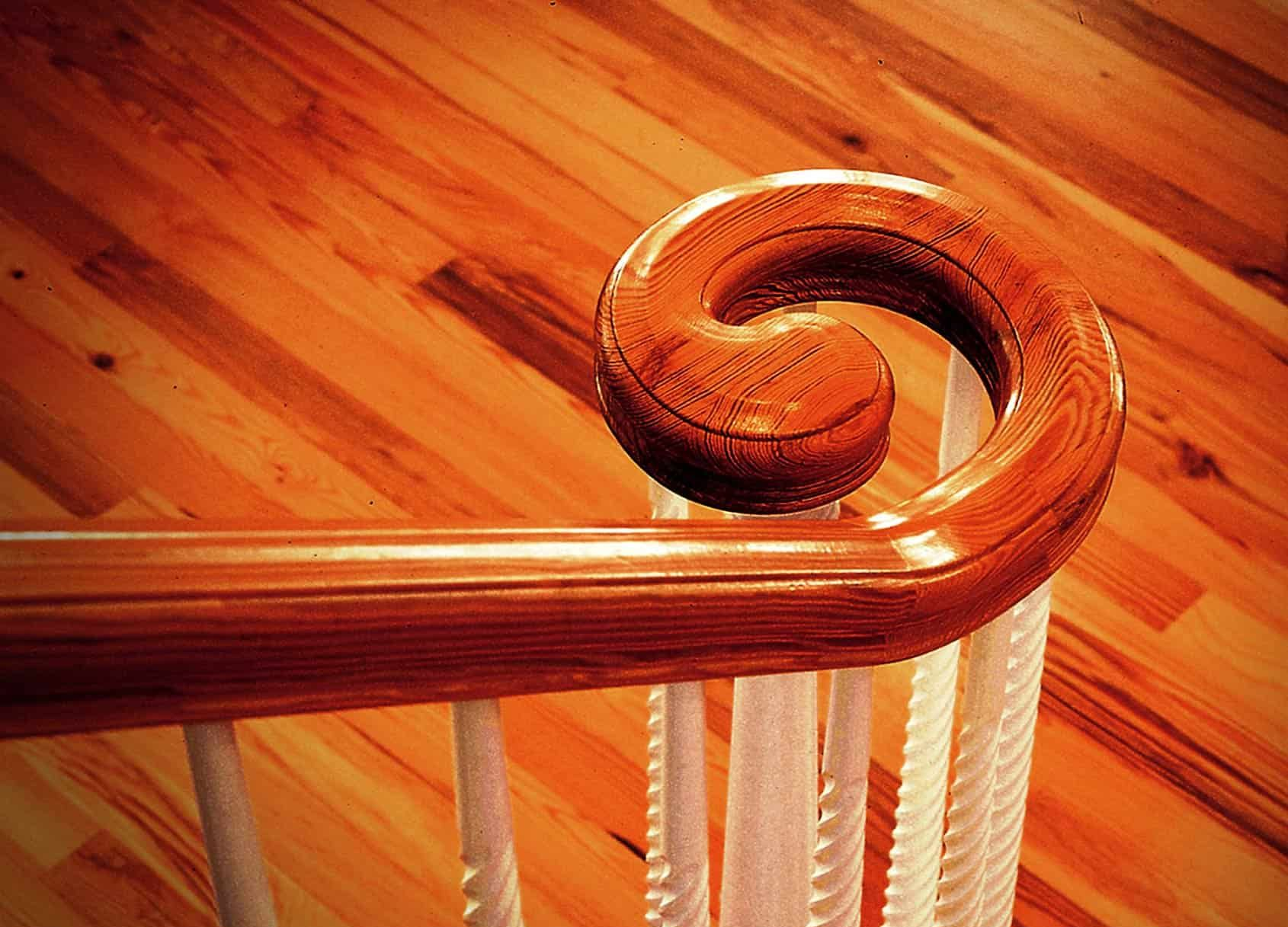 Wooden banister with a scroll detail, set against a backdrop of wooden flooring and white spindles.