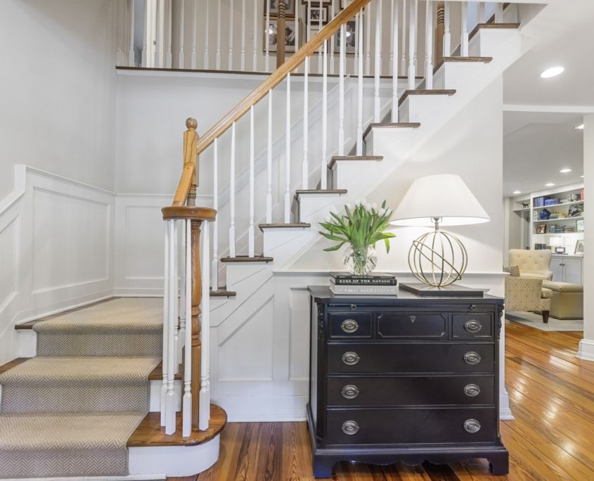 Entryway with staircase, chest of drawers, lamp, and vase of flowers against a white wall and hardwood floor.