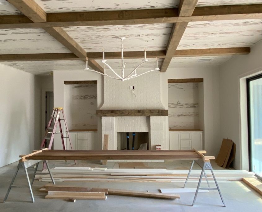 Interior of a room under construction with white walls, exposed beams, fireplace, and a light fixture.
