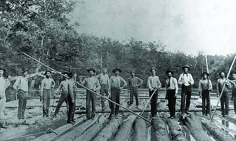 Men standing on logs, holding long poles, likely working on a river or lake, outdoors.