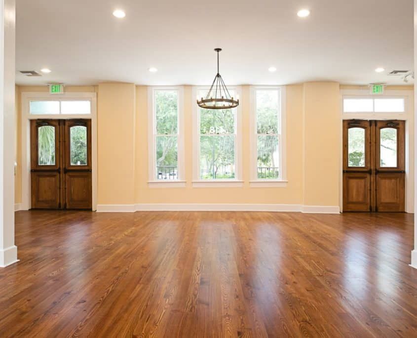 Empty ballroom with wooden floor, tan walls, and a chandelier. Two doors and windows with natural light.