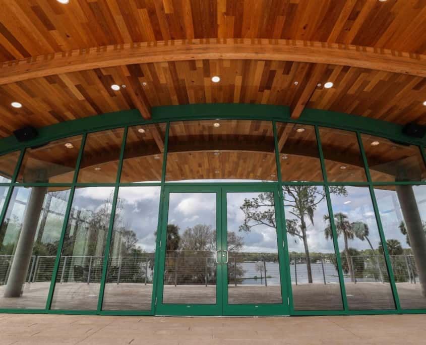 Glass-walled building with a green frame, wooden ceiling, and a view of a lake and trees.