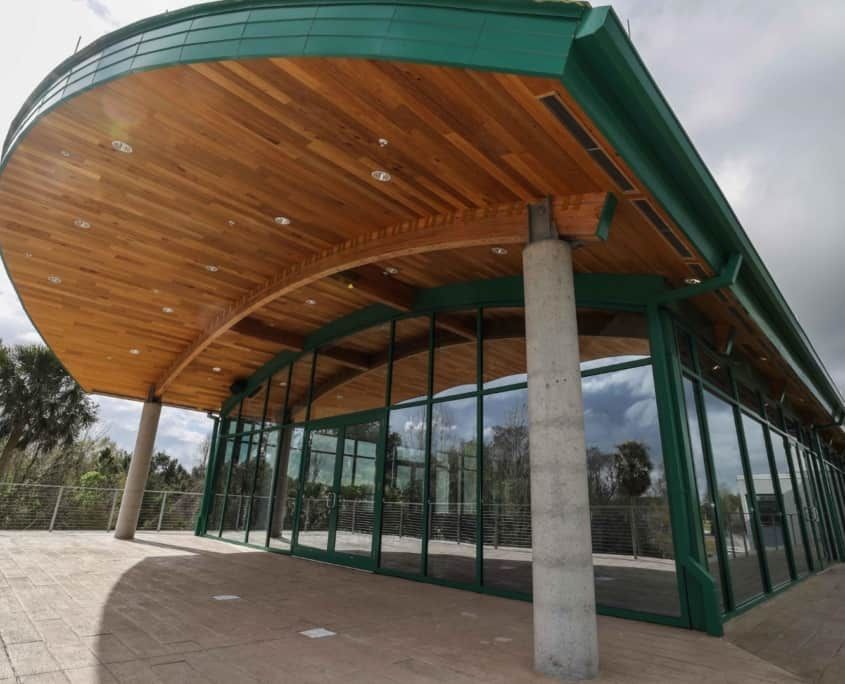 A modern pavilion with glass walls and a curved green roof, wooden ceiling, supported by concrete columns.
