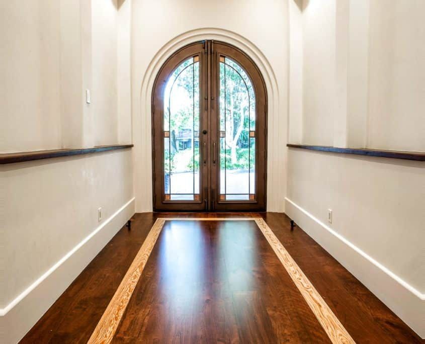 Hallway with arched wooden double doors leading outside, framed by light walls and dark wood floor.
