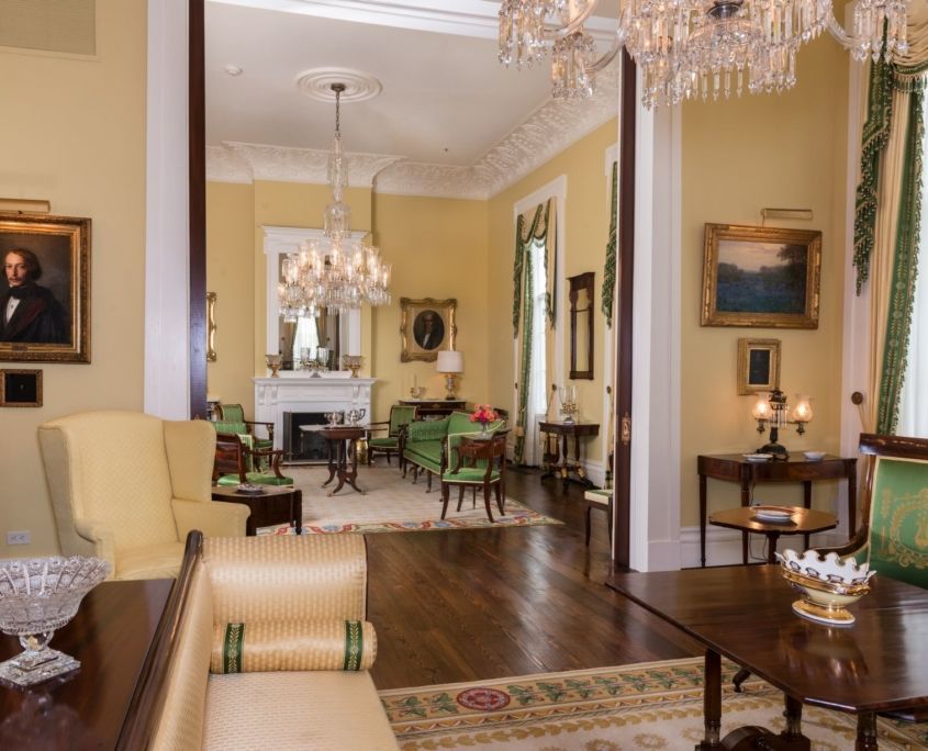Elegant, yellow-walled room with chandeliers, green furniture, paintings, and dark wood floors.