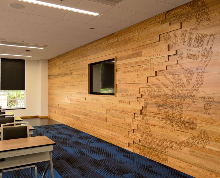 Classroom with wood-paneled wall, embedded monitor, and abstract image. Blue carpet, tables, and a window with blinds.