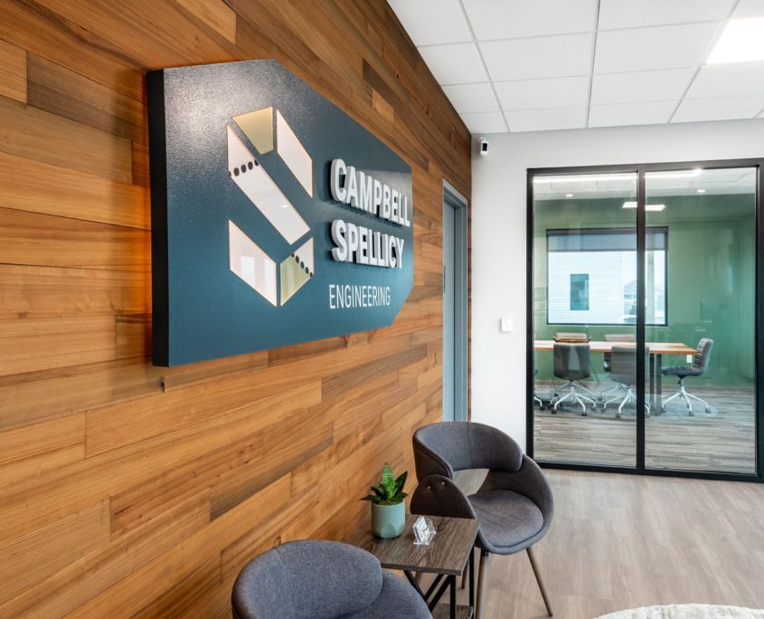 Office lobby with company sign, chairs, and a glass-walled conference room. Wooden wall panels are visible.