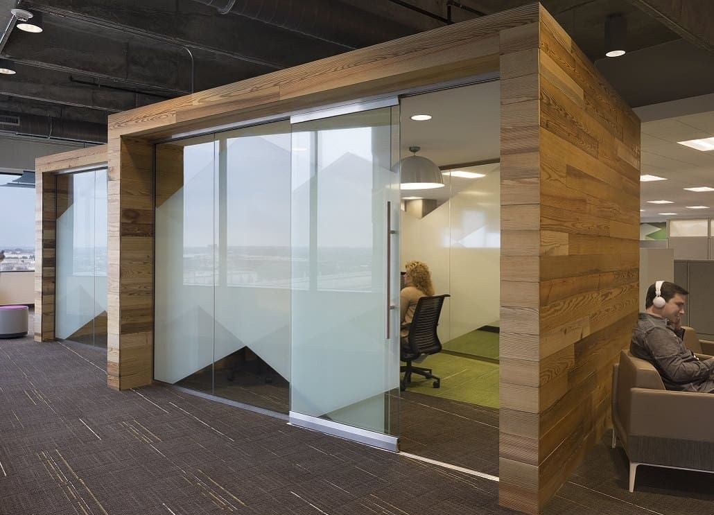 Wooden-framed glass cubicles with frosted sliding doors in an office setting. One person works inside a cubicle; another sits outside.