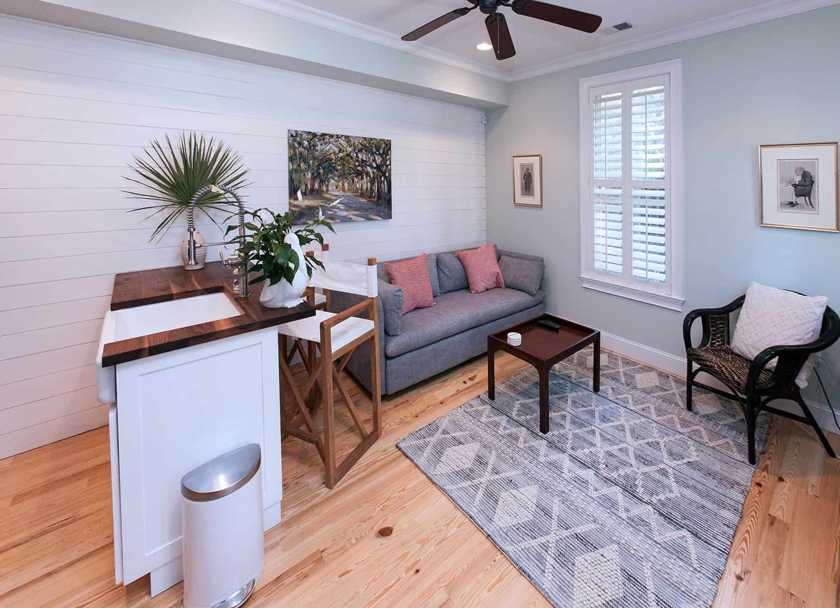 Small, light-filled living space with white panel wall, small couch, kitchen bar, rug, and wicker chair.